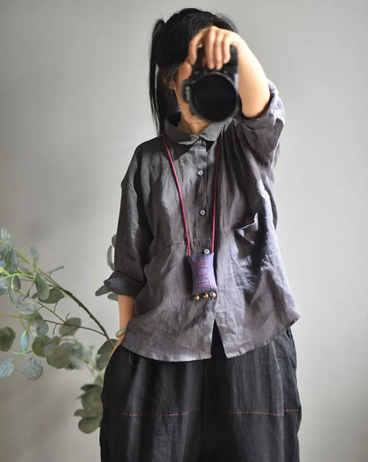 A person wearing a dark gray linen button-up shirt and black pants, holding a black camera in front of their face. They are wearing a purple fabric pouch with a red cord and small gold bells around their neck.