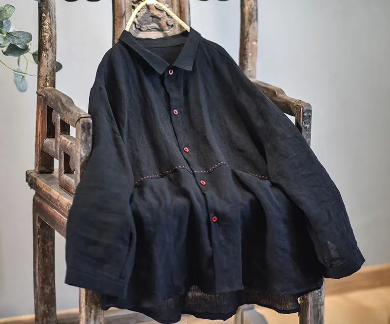 A loose-fitting black linen button-up shirt with red buttons and decorative red stitching, hanging on a wooden chair.