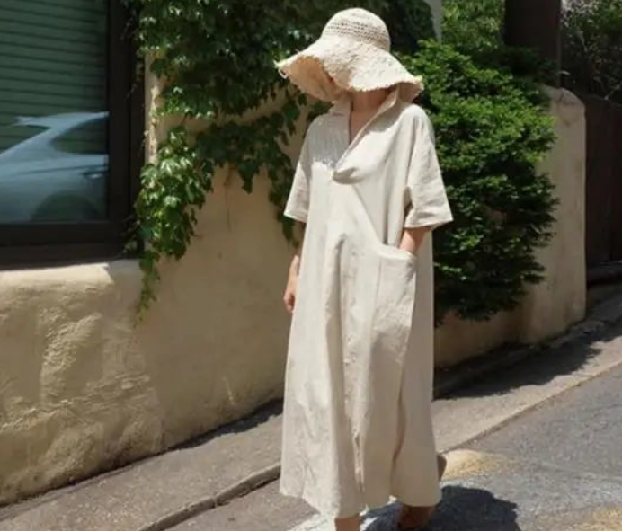 Person wearing a beige dress and hat standing on a street with greenery in the background
