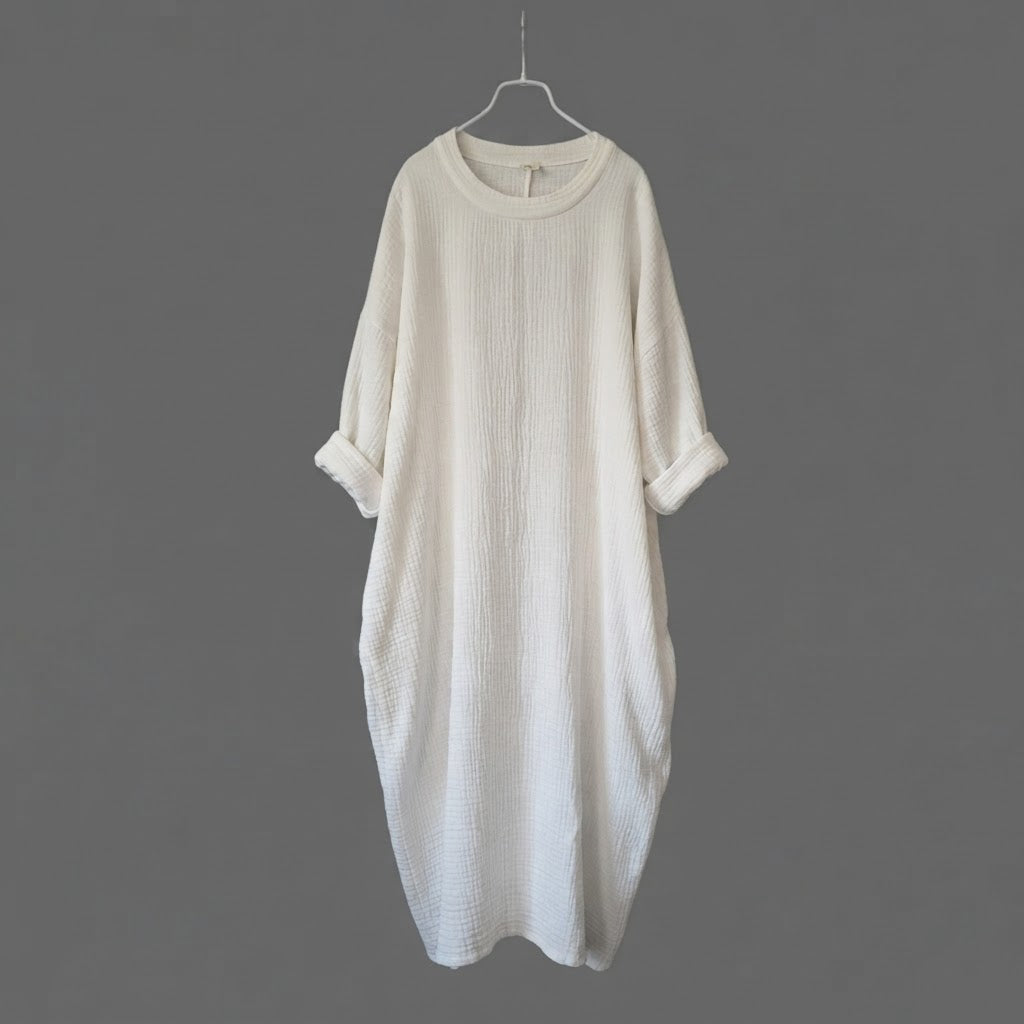An off-white textured cotton dress with three-quarter sleeves and rolled cuffs, hanging on a metal hanger against a solid dark gray background.