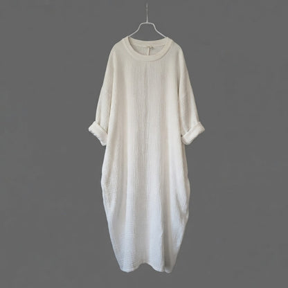An off-white textured cotton dress with three-quarter sleeves and rolled cuffs, hanging on a metal hanger against a solid dark gray background.