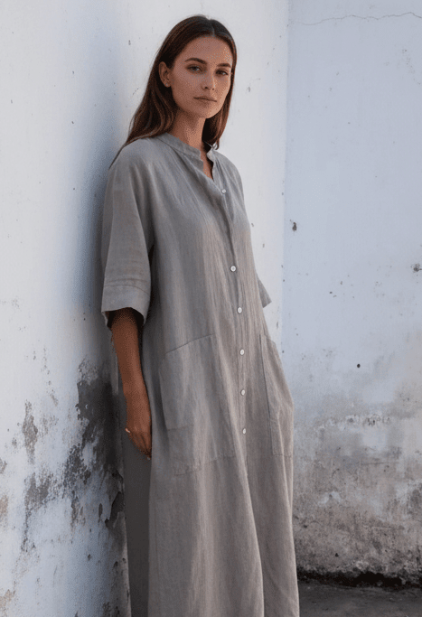 A woman is wearing a long linen dress with a band collar, raglan sleeves, button up.