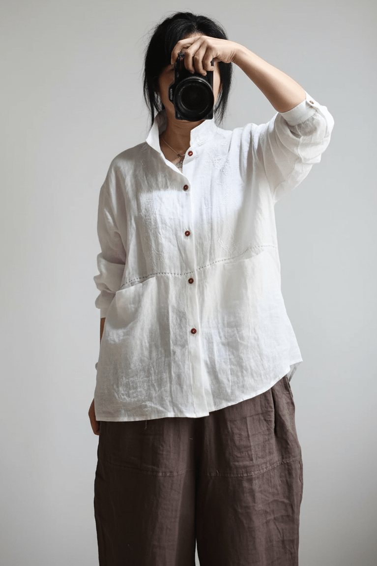 A person wearing a loose white linen button-up shirt and dark brown linen pants, holding a black camera in front of their face against a plain light gray background.