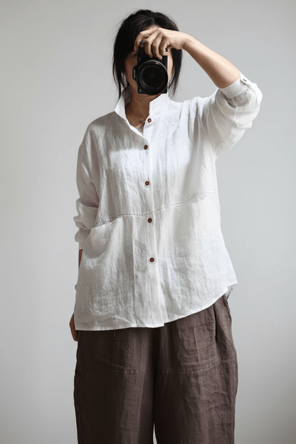 A person wearing a loose white linen button-up shirt and dark brown linen pants, holding a black camera in front of their face against a plain light gray background.