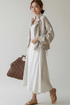 A woman models a light beige linen jacket over a white midi dress, holding a brown woven tote bag against a plain light gray studio background.