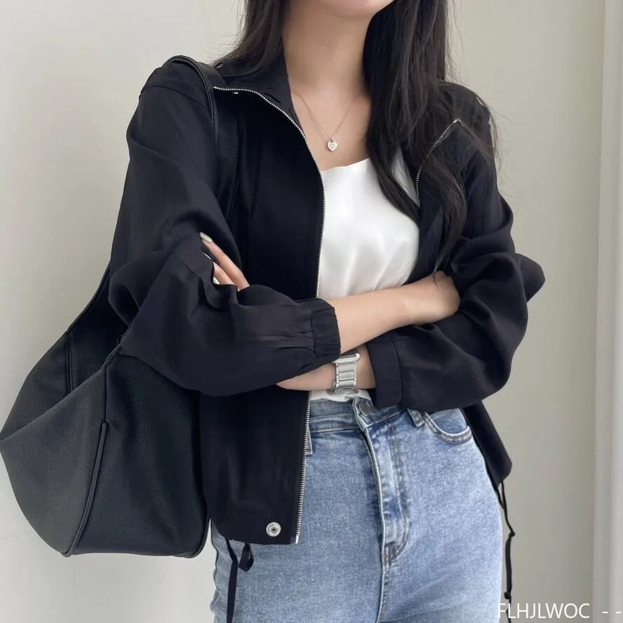 Person wearing a black jacket over a white top and blue jeans against a neutral background