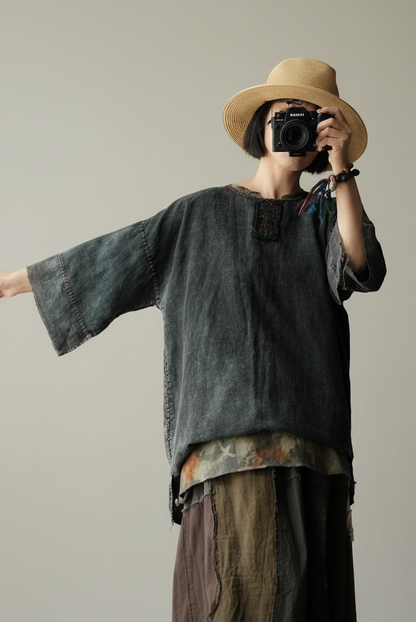 A person wearing a wide-brimmed straw hat, a dark washed short-sleeve top, and a patchwork skirt holds a camera in front of their face against a plain light gray studio background.