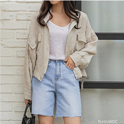 Person wearing a beige jacket, white shirt, and denim shorts against a stone wall.