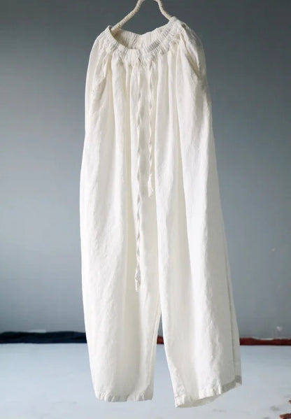 A pair of loose, off-white linen pants hanging from a rope hanger against a plain gray wall.