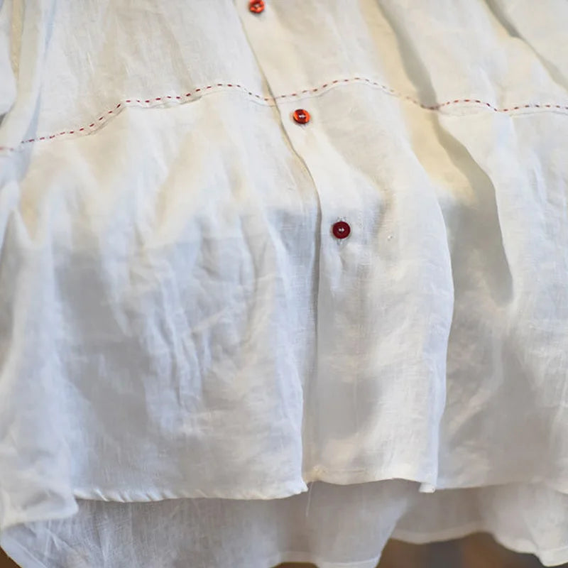 Close-up of a white textured fabric garment, likely a linen shirt or blouse, featuring red buttons and red dashed contrast stitching.