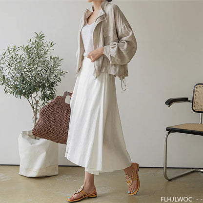 Person wearing a beige jacket and white pants in a minimalistic room with a plant and chair.