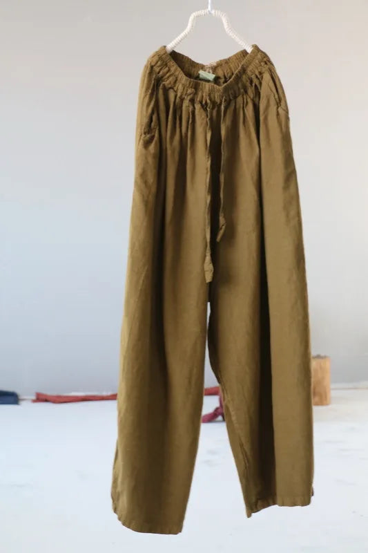 A pair of loose, wide-leg trousers in an olive-brown or khaki shade, hanging from a white plastic hanger against a plain light gray wall.