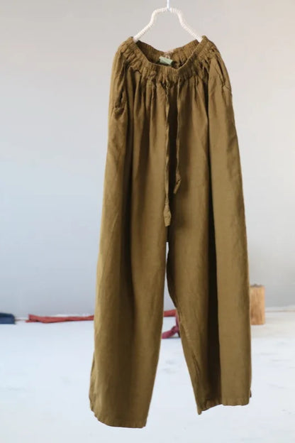 A pair of loose, wide-leg trousers in an olive-brown or khaki shade, hanging from a white plastic hanger against a plain light gray wall.