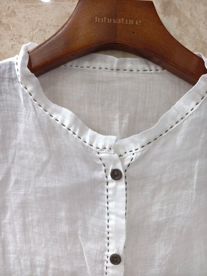 a loose white long-sleeve blouse with gathered cuffs and a button placket, paired with a dark skirt, photographed in a dim indoor setting.