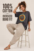 A woman models an oversized dark gray t-shirt with a retro 1970s-style circular graphic, paired with beige jogger pants and sandals, sitting on a white stool against a light beige background. Text on the left reads &