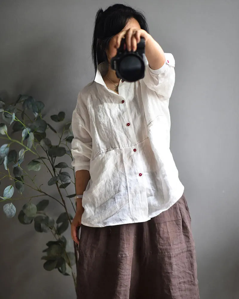 A person wearing a loose white button-up shirt with small red buttons and a brown skirt, holding a black camera in front of their face against a gray wall with a green plant on the left.
