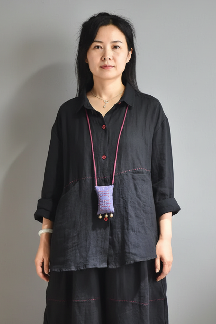 A woman wearing a loose black linen button-up shirt with rolled sleeves and matching black pants, standing against a plain gray background.