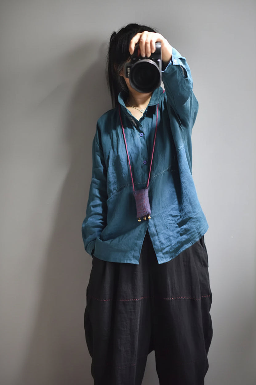 A person wearing a blue button-up shirt and black pants, holding a camera in front of their face. A small purple pouch hangs from the shirt&