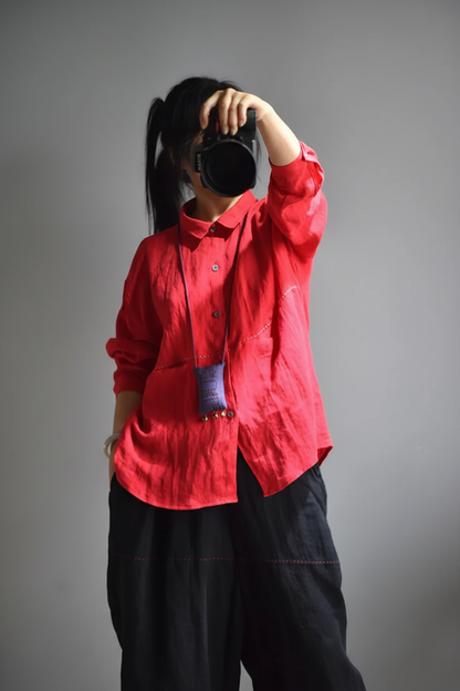 A person wearing a bright red button-up shirt and loose black pants, holding a black camera in front of their face against a plain gray studio background.