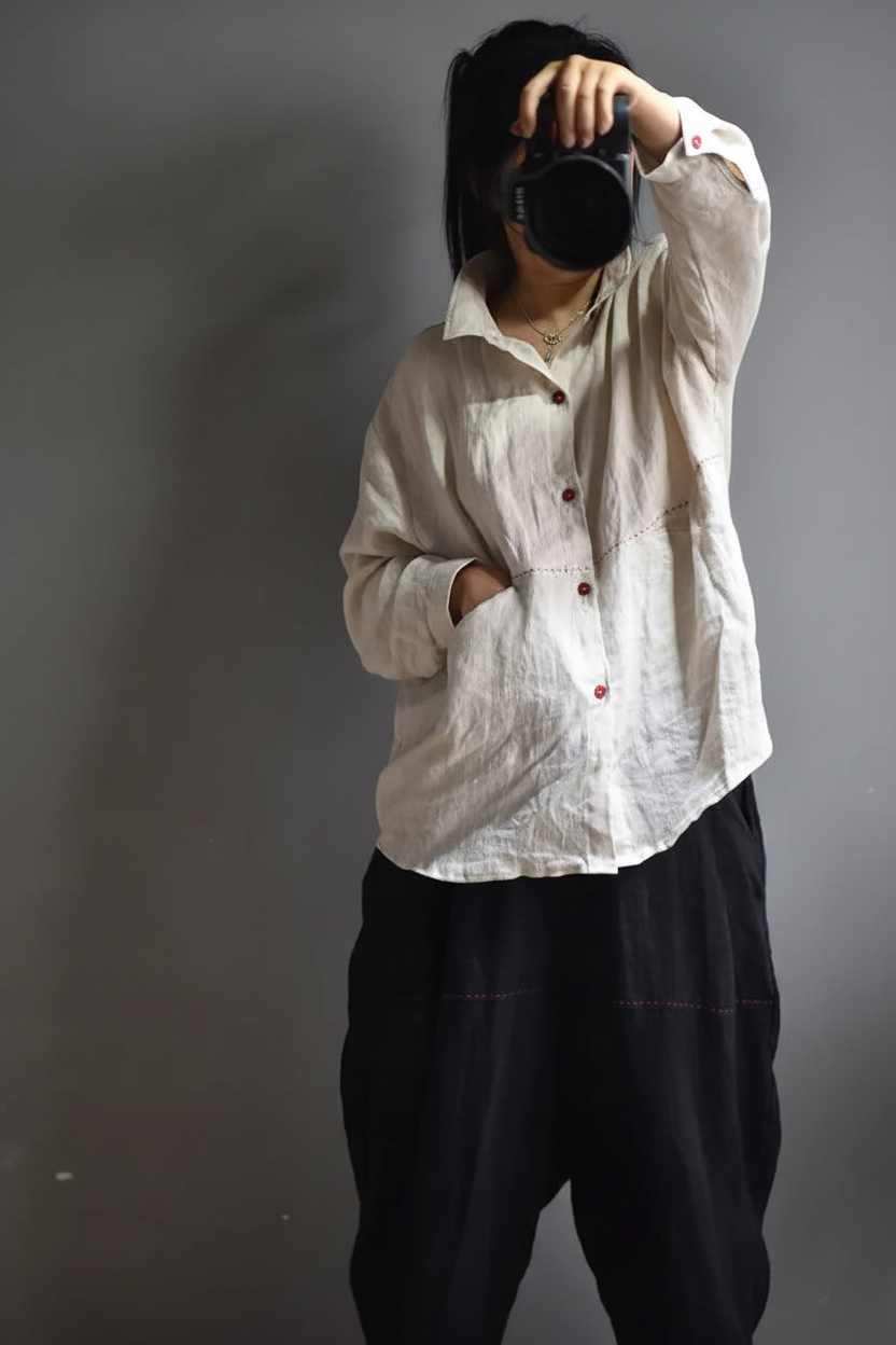 A person wearing a loose, off-white button-up shirt and dark baggy pants, holding a black camera to their face against a plain gray background.