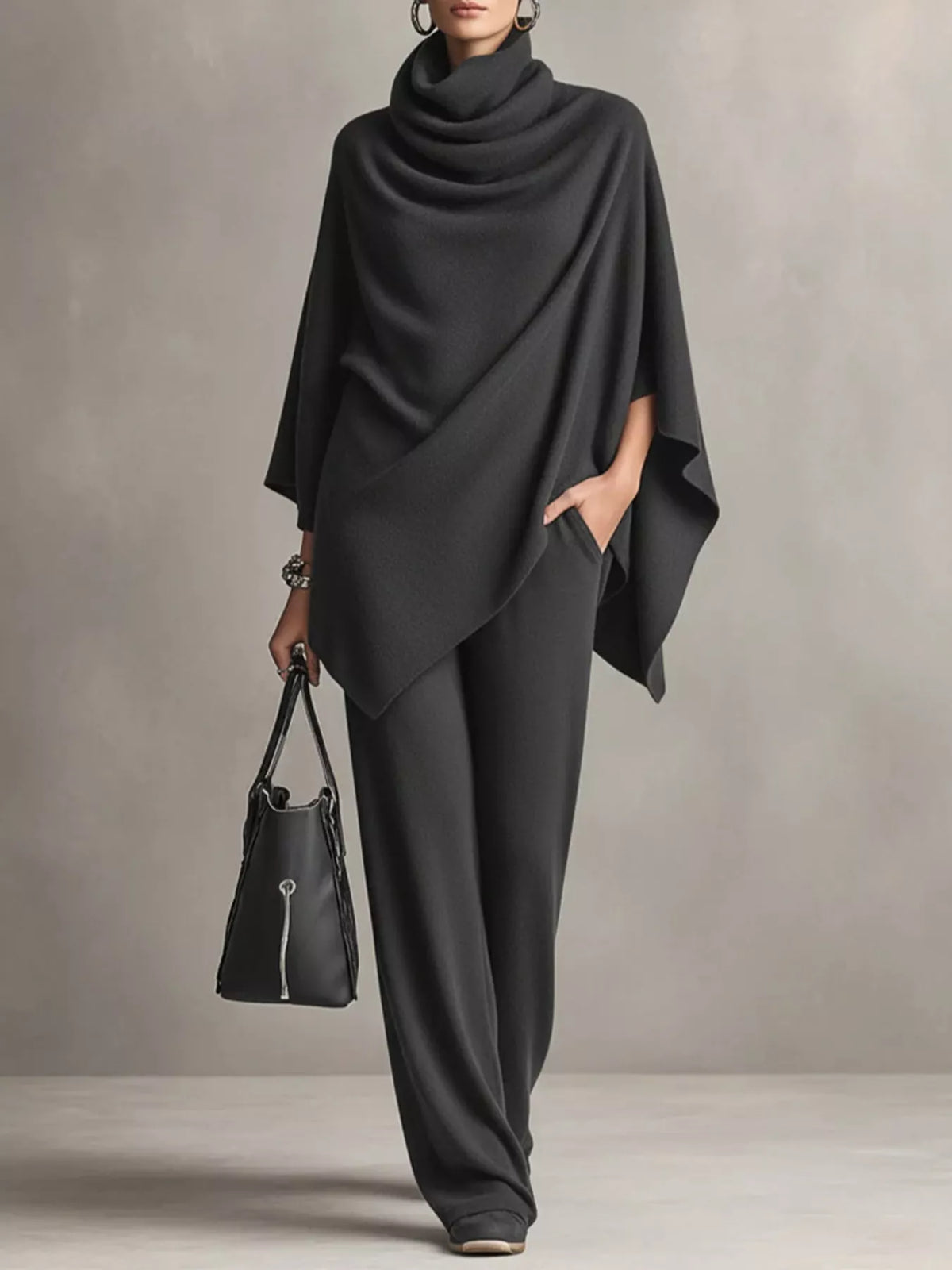 A model wearing a dark charcoal gray draped poncho top and matching wide-leg trousers, holding a black handbag against a neutral gray studio background.