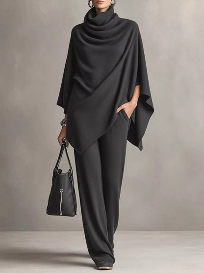 A model wearing a dark charcoal gray draped poncho top and matching wide-leg trousers, holding a black handbag against a neutral gray studio background.