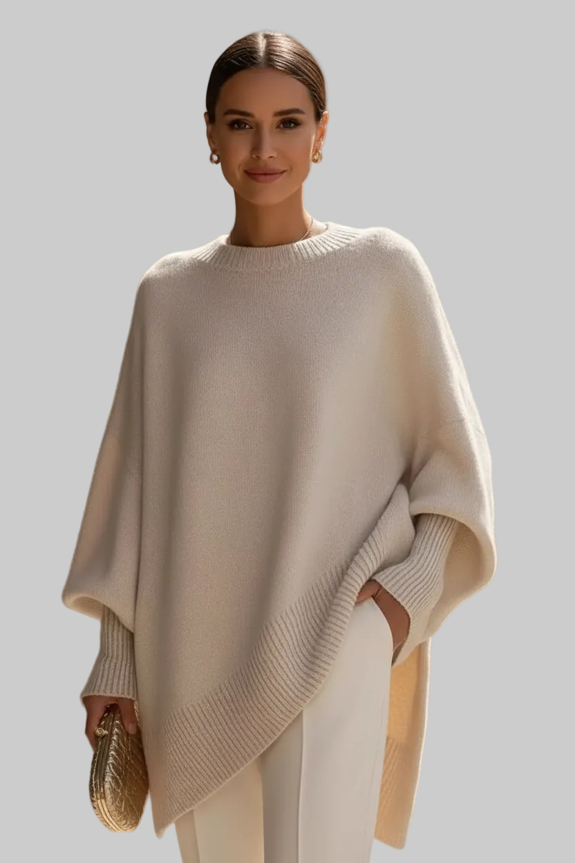 Woman wearing a beige poncho and holding a gold clutch against a neutral background