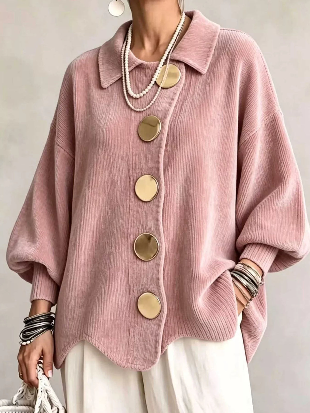 A person wearing an oversized dusty pink corduroy-style knit cardigan with large round gold-tone buttons, styled with layered pearl necklaces, bracelets, and white pants against a soft neutral background.