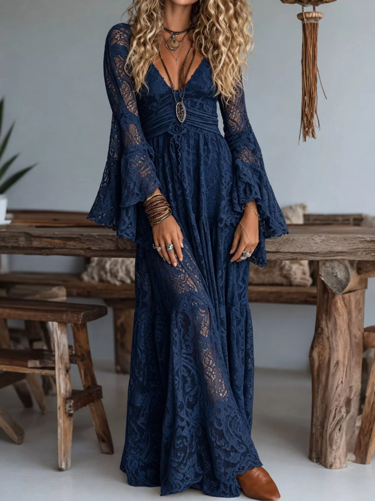 A woman wearing a navy blue lace maxi dress with long flared sleeves, a deep V-neckline, and a drawstring waist, standing in a rustic indoor setting with wooden furniture.