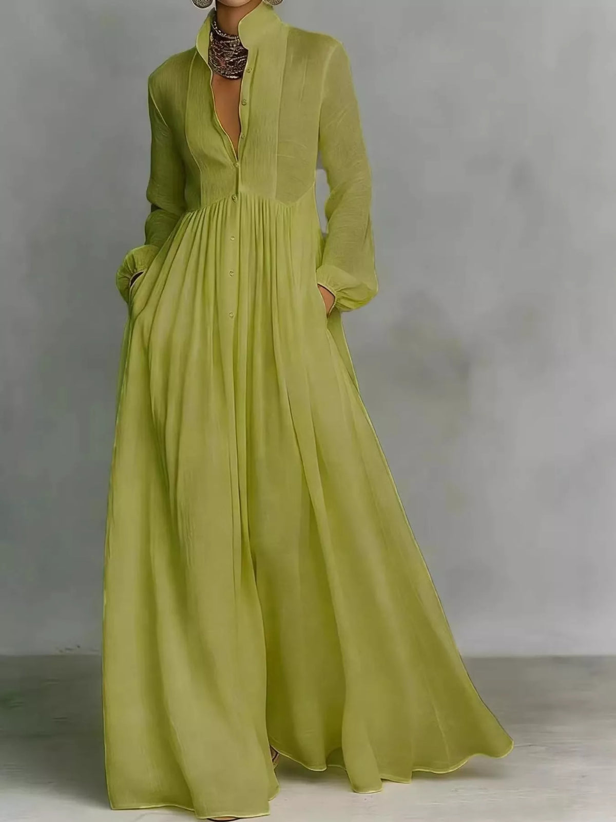 A woman wearing a flowing maxi dress in a light olive green color, standing against a neutral gray studio background.