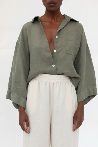 A model wearing an olive green linen button-down shirt with wide sleeves and white linen trousers, shot against a plain white background.