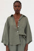 A model wearing an olive green linen button-down shirt with wide sleeves and matching trousers, photographed against a plain white studio background.
