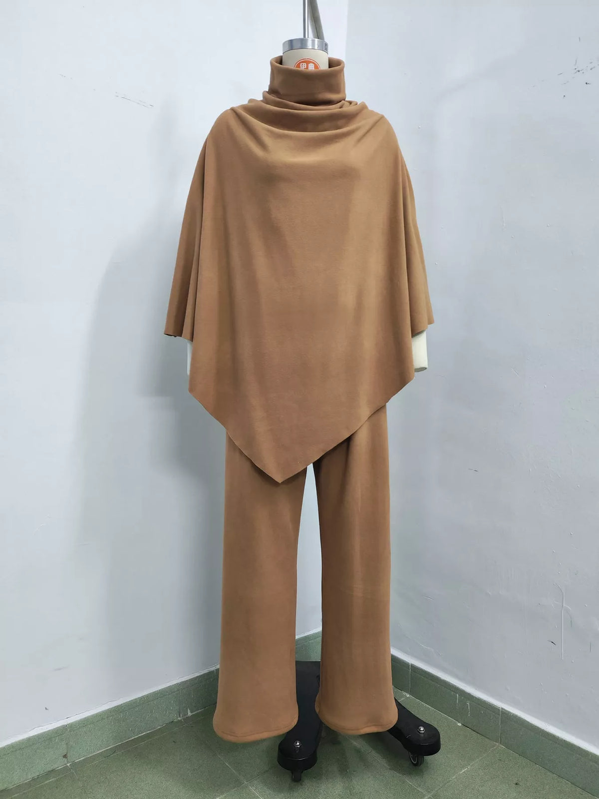 A two-piece clothing set in a solid camel brown color, consisting of a draped turtleneck top and matching wide-leg pants, displayed on a mannequin in a corner room.