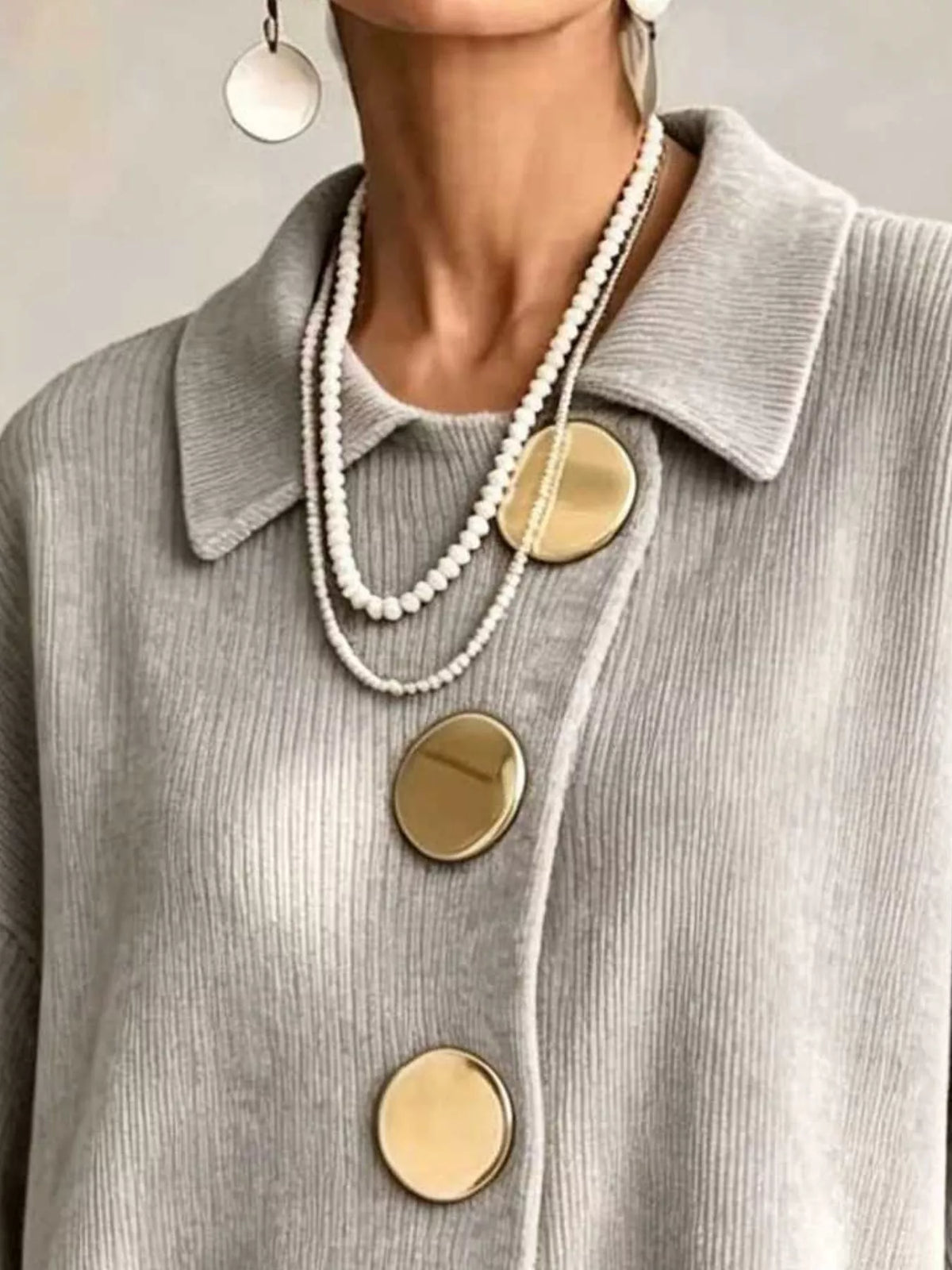 Close-up of a person wearing a light gray ribbed knit collared top with large round gold buttons, layered with a long white pearl necklace and gold disc earrings.