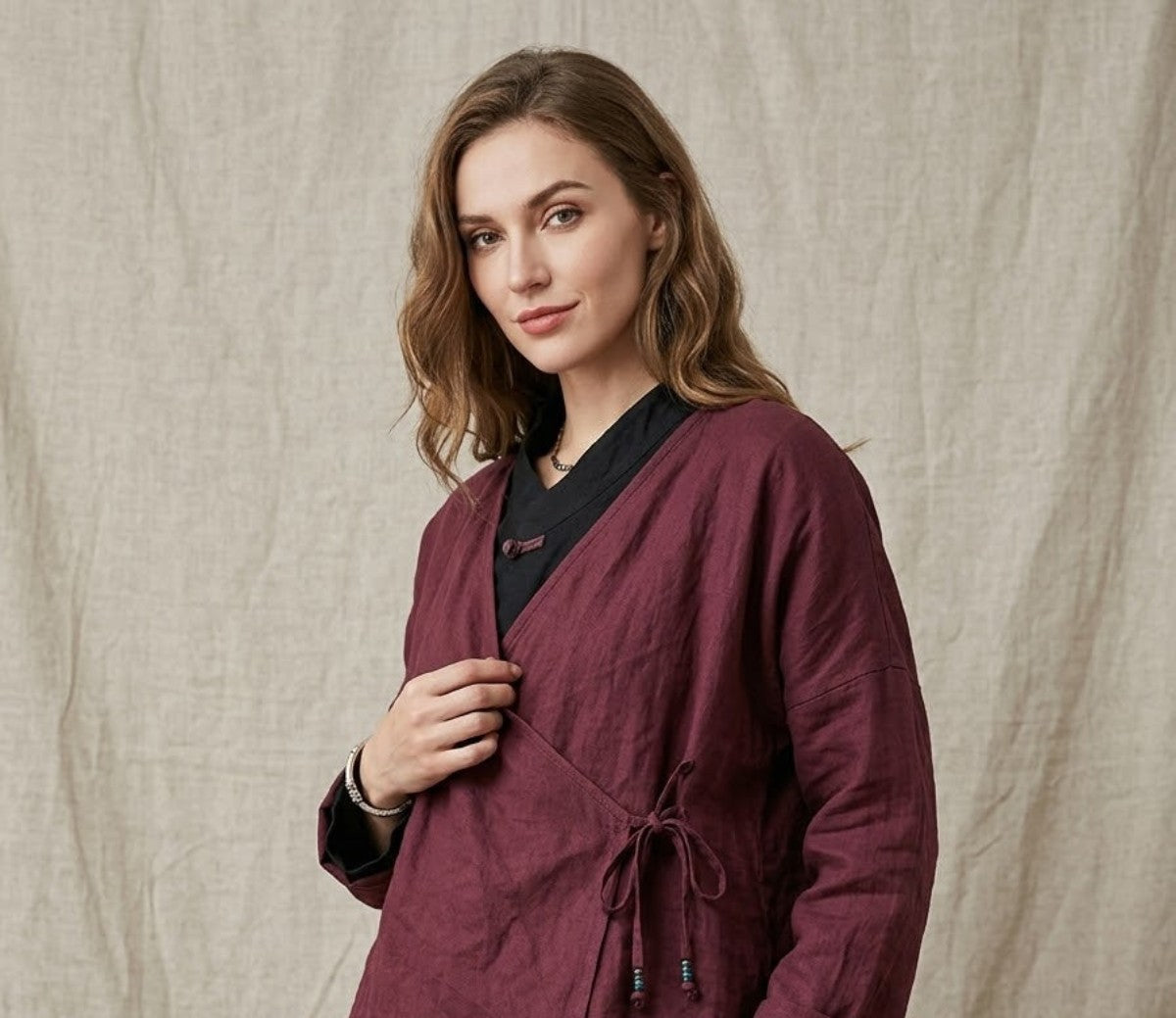 A woman wearing a burgundy wrap-style linen jacket with a drawstring waist over a black top and black skirt, standing against a beige linen backdrop.