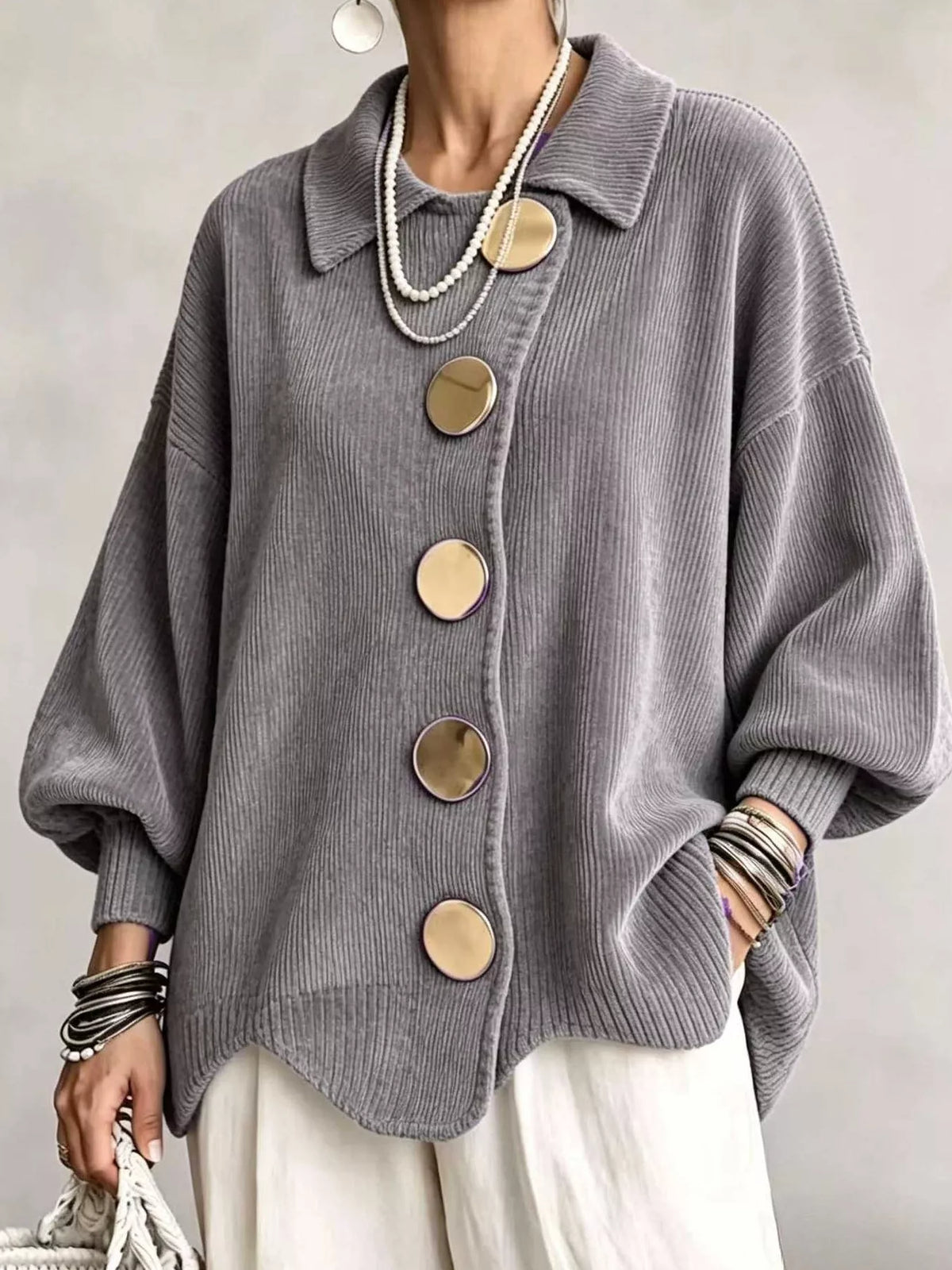 A person wearing an oversized gray ribbed knit cardigan with large round gold-tone buttons, paired with white pants and layered necklaces, against a plain light gray background.