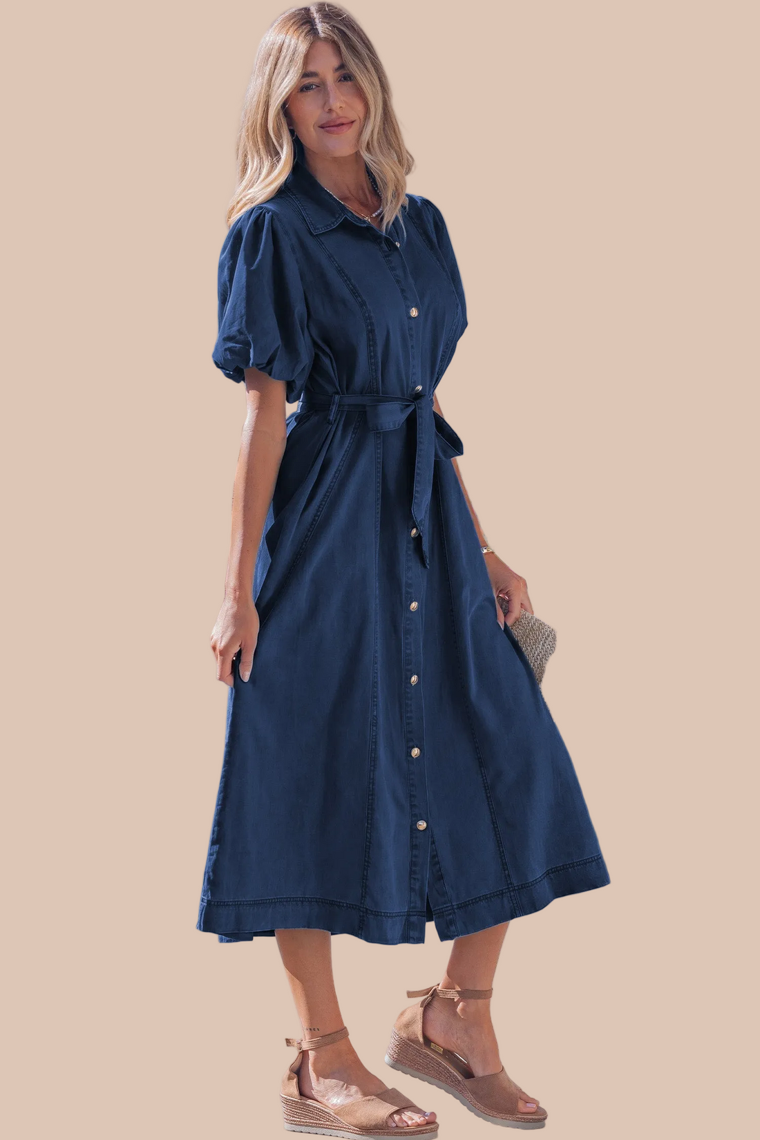 A woman modeling a dark blue denim midi shirt dress with puff sleeves, a button-down front, and a matching tie belt, set against a solid beige background.
