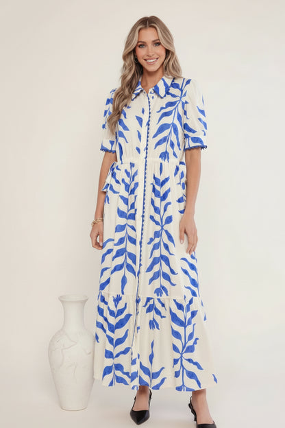 A woman wearing a blue and white patterned dress standing outdoors with plants in the background