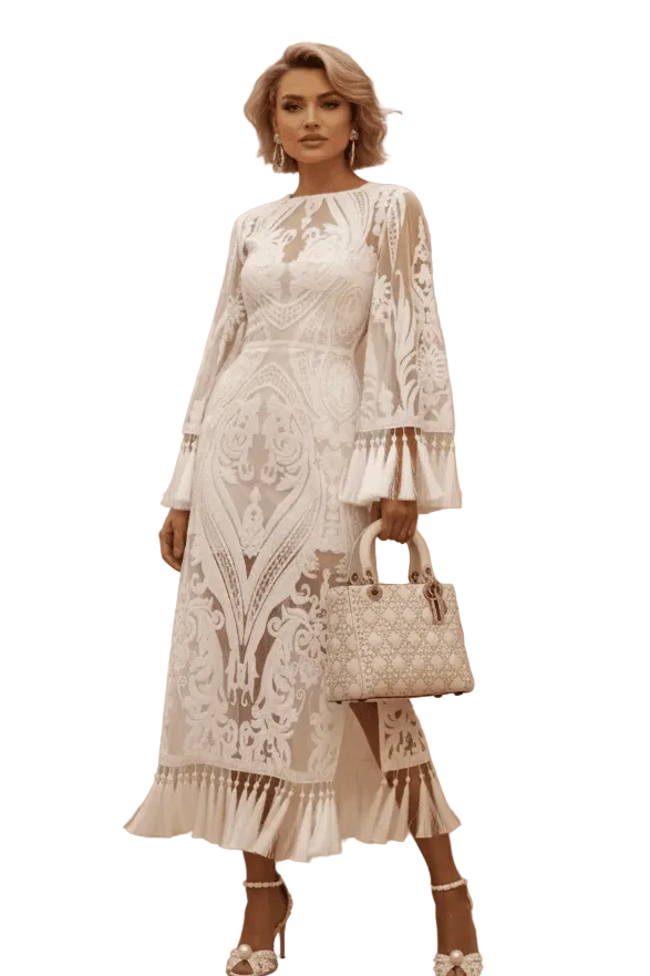 A woman stands in a studio wearing a long-sleeve white lace midi dress with a keyhole neckline and ruffled hem, holding a matching white quilted handbag. The background is a warm beige wall with a wooden floor and a large vase of dried pampas grass.
