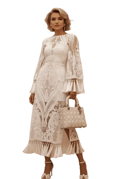 A woman stands in a studio wearing a long-sleeve white lace midi dress with a keyhole neckline and ruffled hem, holding a matching white quilted handbag. The background is a warm beige wall with a wooden floor and a large vase of dried pampas grass.