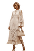 A woman stands in a studio wearing a long-sleeve white lace midi dress with a keyhole neckline and ruffled hem, holding a matching white quilted handbag. The background is a warm beige wall with a wooden floor and a large vase of dried pampas grass.