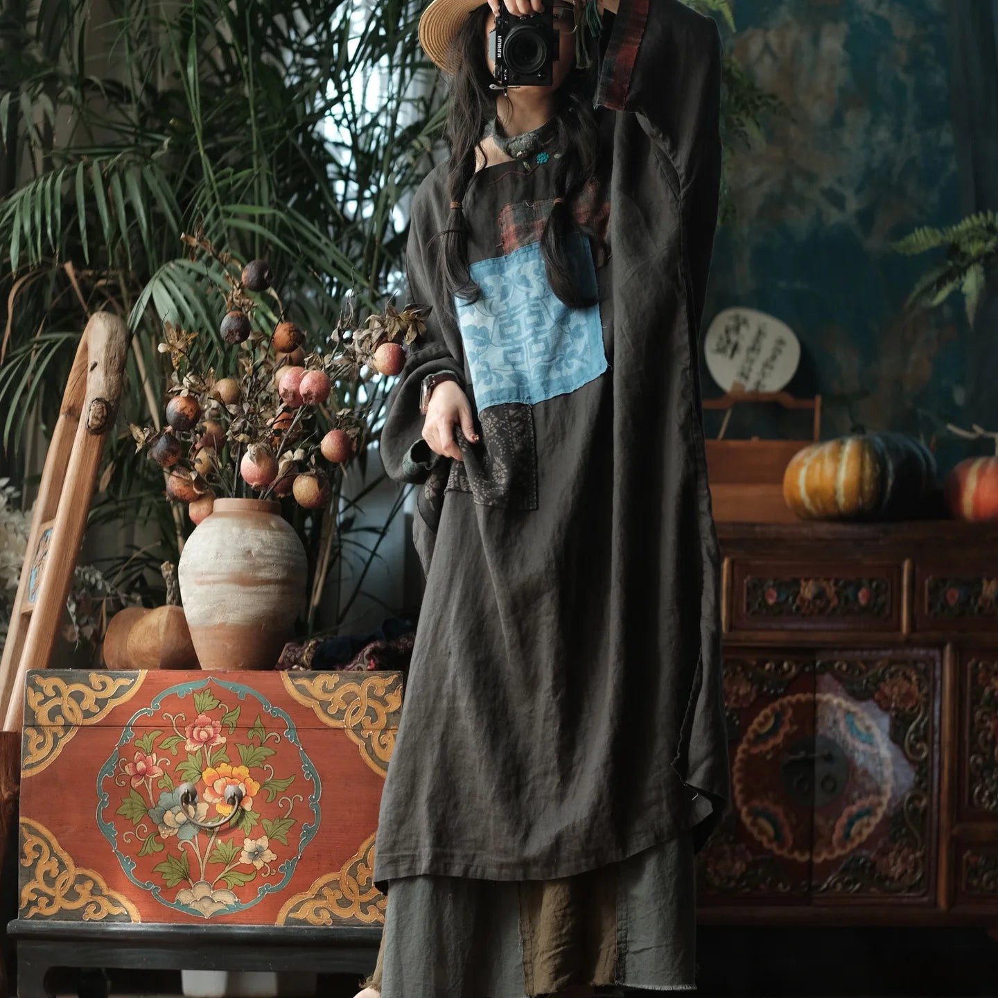 A person wearing a dark brown patchwork linen dress with a light blue square patch and a brown hat, holding a camera in a rustic, decorated room with plants and wooden furniture.