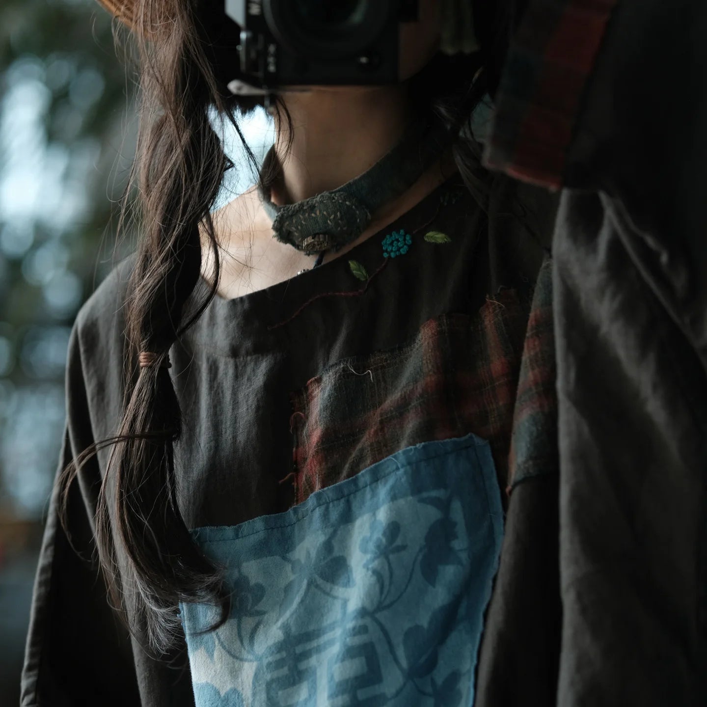 A close-up lifestyle shot of a person wearing a dark patchwork top with a blue floral apron-style panel, a red plaid patch, and a dark textured collar, with long dark hair and a camera partially visible.