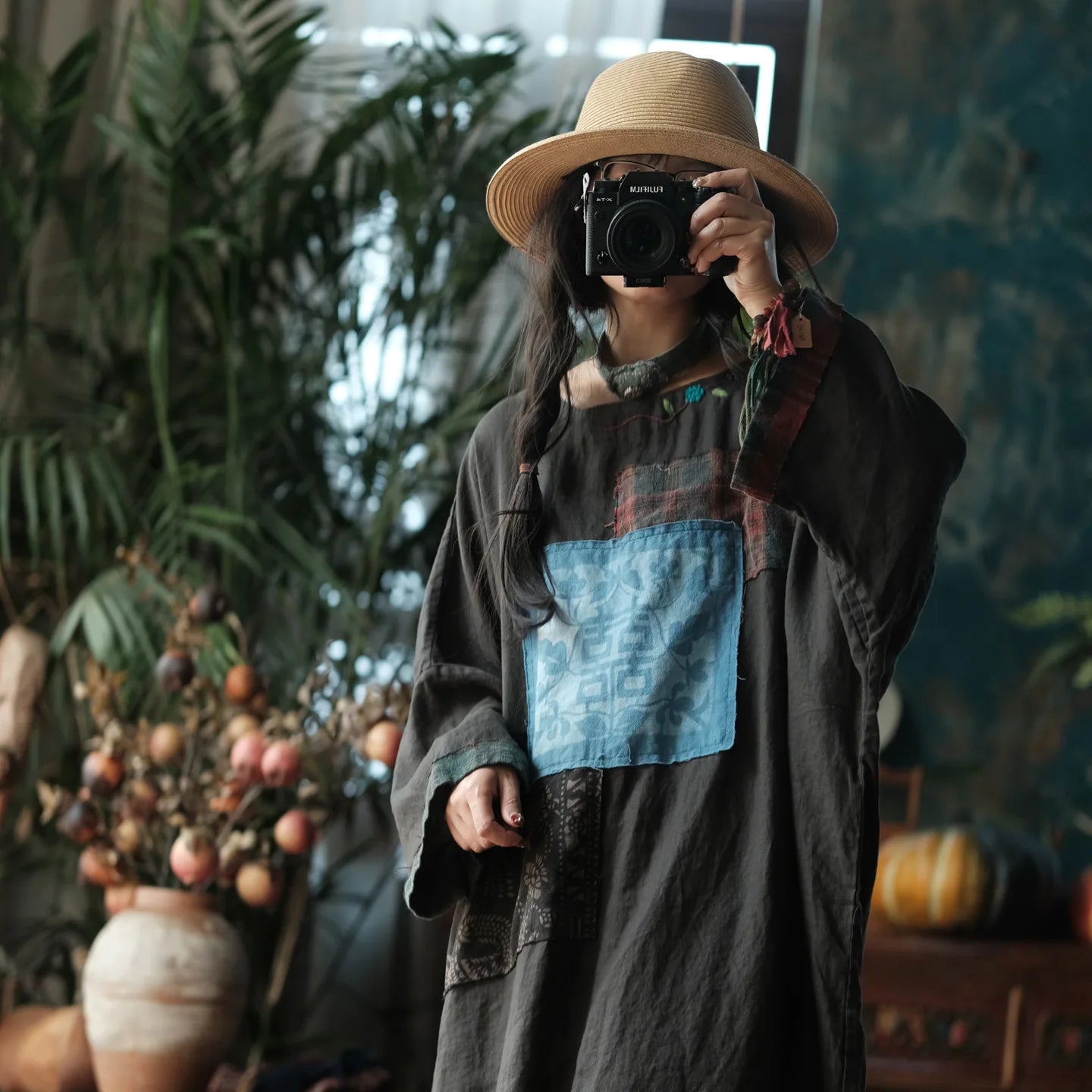 A person wearing a wide-brim straw hat and a dark gray patchwork long-sleeve dress with a light blue square patch on the chest, holding a camera in a mirror selfie inside a decorated room with plants.