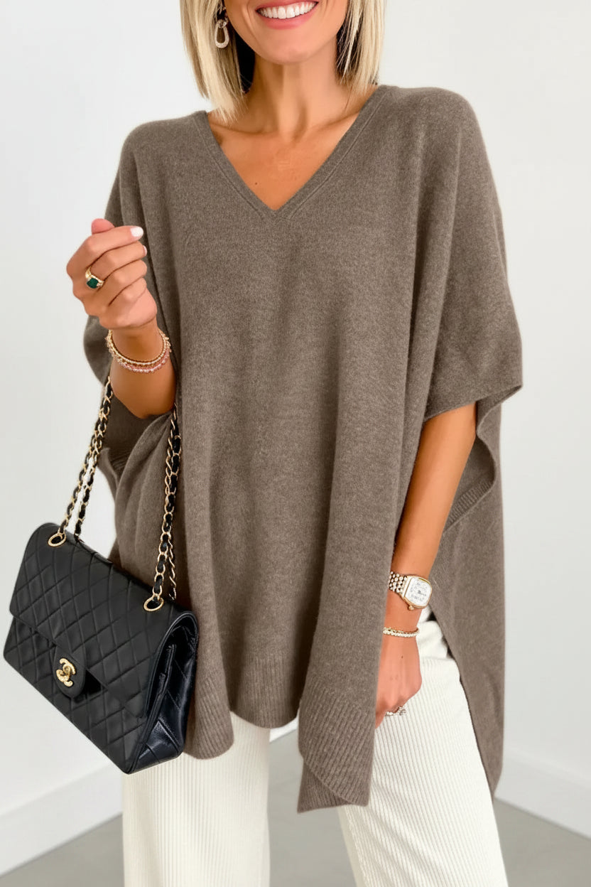 Woman wearing a brown poncho over a blue outfit, holding a black handbag indoors.
