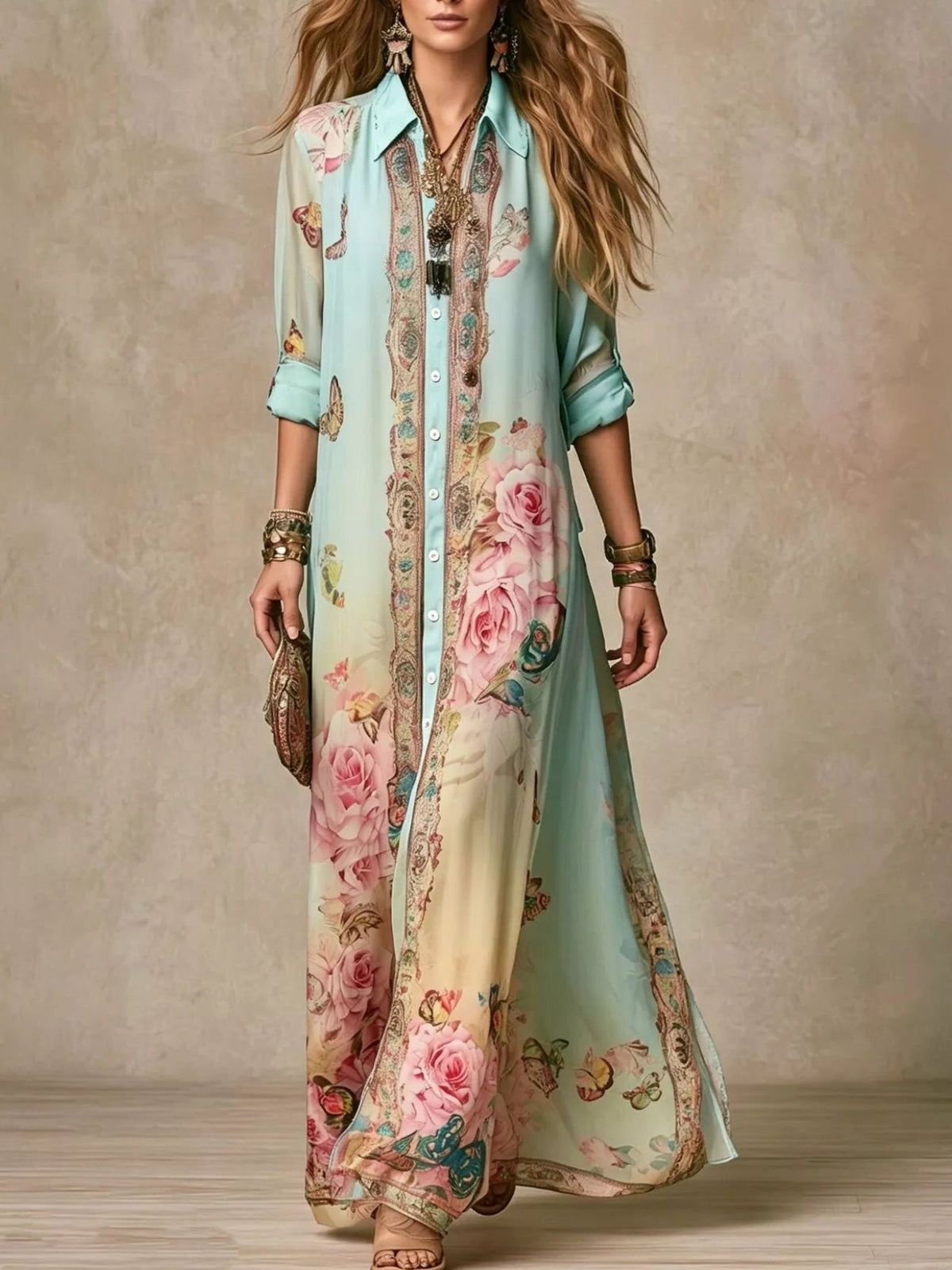 A woman models a long, flowing button-up dress with a light aqua base and a large pink floral print with ornate border detailing, set against a textured beige studio backdrop.
