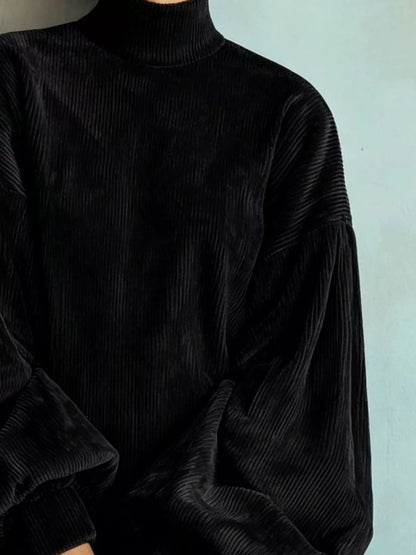 A close-up of a person wearing a black ribbed corduroy mock-neck sweater with voluminous balloon sleeves, set against a pale blue wall.