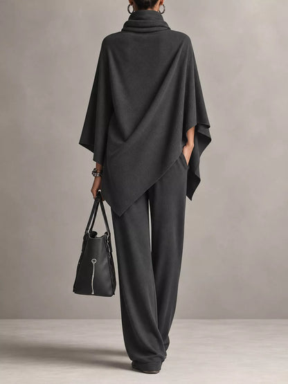 A model from behind wearing a dark charcoal gray cowl-neck poncho and matching wide-leg trousers, holding a black leather handbag against a neutral gray studio background.