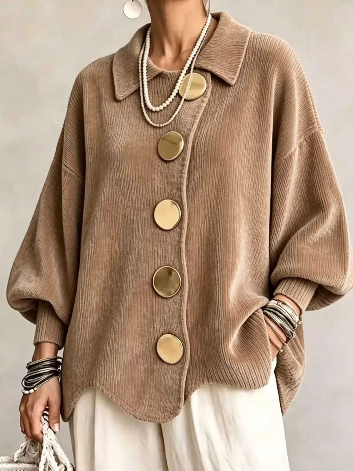 A person wearing an oversized taupe-brown ribbed knit cardigan with large round gold-tone buttons, styled with layered pearl necklaces, bracelets, and white pants against a neutral studio background.