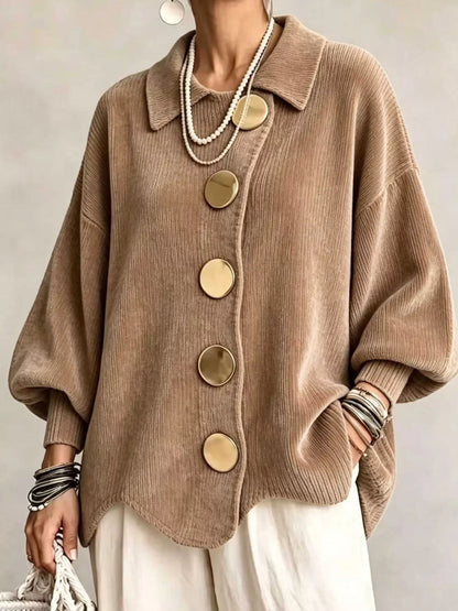 A person wearing an oversized taupe-brown ribbed knit cardigan with large round gold-tone buttons, styled with layered pearl necklaces, bracelets, and white pants against a neutral studio background.